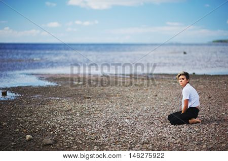 A Young Girl With Short Hair In A White Blouse And Black Pants Is Sitting On His Knees On The Beach