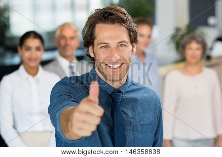 Businessman giving thumb up to camera in front of colleagues. Successful businessman showing thumbs up sign and looking at camera. Young happy manager with business team.