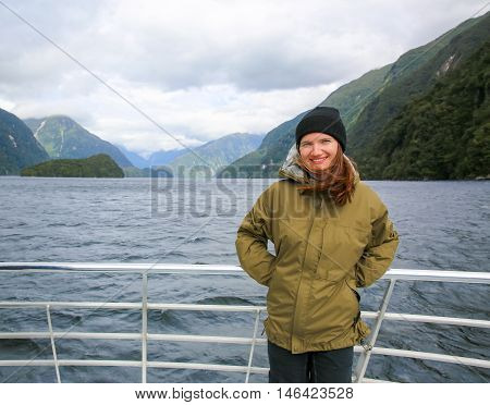 Young smiling woman standing on the deck of touring boat. Picturesque view of Milford Sound at Te Anau. Fiordland National Park New Zealand.