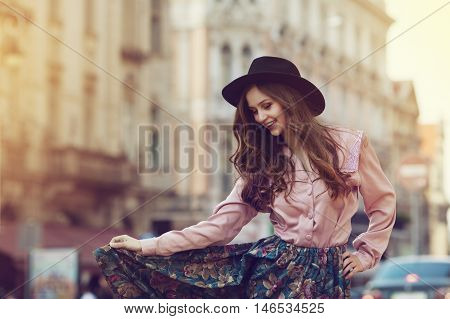Outdoor portrait of young beautiful happy lady posing on street. Model wearing stylish clothes. Girl looking down. Sunny day. Female fashion. City lifestyle. Copy space. Toned style instagram filters.