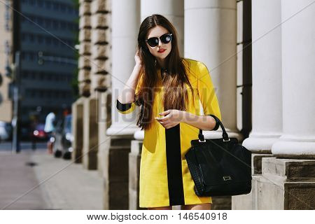 Outdoor portrait of young beautiful lady walking on the street. Model wearing sunglasses and stylish yellow summer dress. Girl looking down. Female fashion concept. City lifestyle. Sunny day. Waist up.