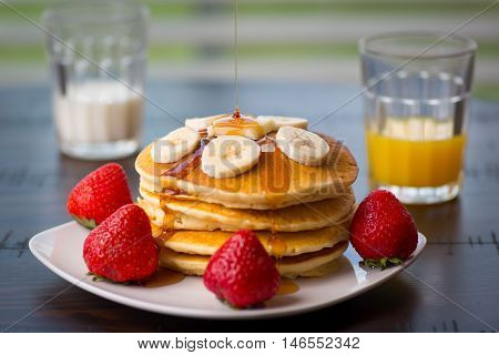 Pancakes whip cream, strawberry, bananas, milk with a glass of orange juice at the kitchen table, with syrup pouring on it.