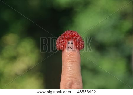 Red raspberry on the thumb with a sad face in front of green nature