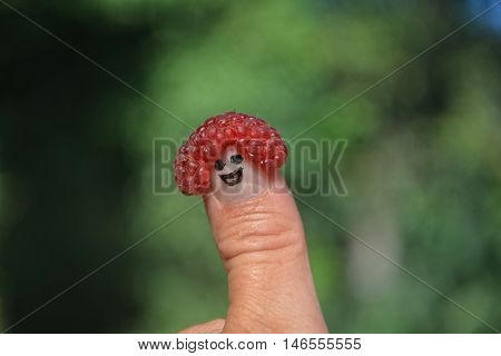 Red raspberry on the thumb with a smiling face in front of green nature