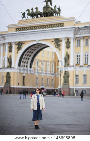 Young beautiful girl student in a blue denim dress and white jacket standing on Palace Square near the Arc de Triomphe of the Head of Staff in St. Petersburg