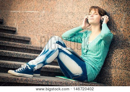 Young woman listening music in Headphones. Beautiful young female student.  Woman sitting on stairs in city park.