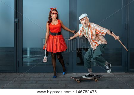 Old man with a cane to skateboard next to a young girl.
