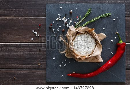 Cheese delikatessen closeup with chili pepper on black stone surface and wood. Camembert or brie circle in brown kraft paper decorated with basil, top view image