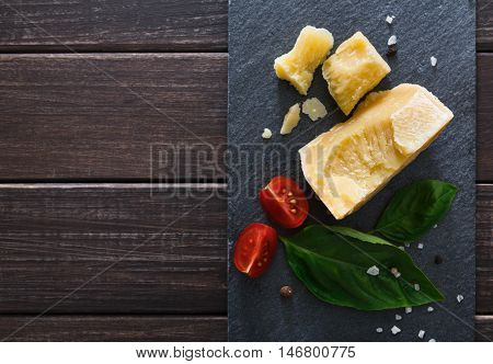 Cheese delikatessen closeup on black stone desk at wooden surface background. Parmesan pieces decorated with basil and cherry tomatoes, top view image with copy space