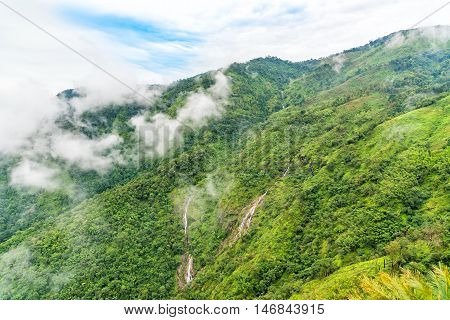 Small waterfall on green mountain ridge in rain forest on cloudy day.