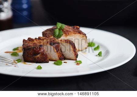 Korean style sliced glazed pork chops on white plate with sticky citrus caramelized sauce with oranges and mint leaves. Salt, pepper, knife and green mint leaves near pan with chops.