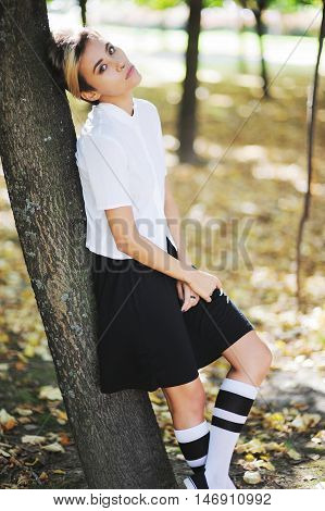 Seriously, young teen girl in a school uniform is resting at recess, leaning against a tree in autumn park and looking pensively.