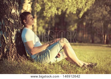 Relaxing man sitting under a tree with eyes closed meditating and enjoying the warm evening sunset in profile