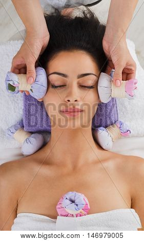 Herbal ball face massage in ayurveda spa. Female massagist with young woman in wellness center. Healthcare therapy to beautiful indian girl in beauty parlor, top view of face with eyes closed