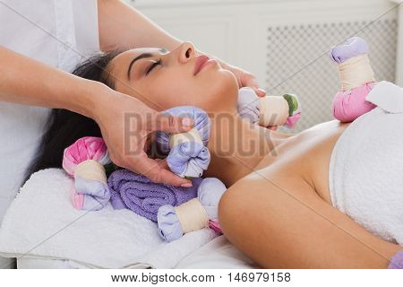 Herbal ball face massage in ayurveda spa. Female massagist with young woman in wellness center. Healthcare therapy to beautiful indian girl in beauty parlor, side view of face with eyes closed