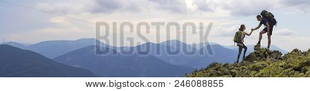 Young Tourists With Backpacks, Athletic Boy Helps Slim Girl To Clime Rocky Mountain Top Against Brig