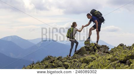 Young Tourists With Backpacks, Athletic Boy Helps Slim Girl To Clime Rocky Mountain Top Against Brig