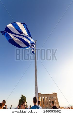 Greek Flag Waving On A Mast At The Acropolis Surrounded By Tourists With The Parthenon Visible In Th