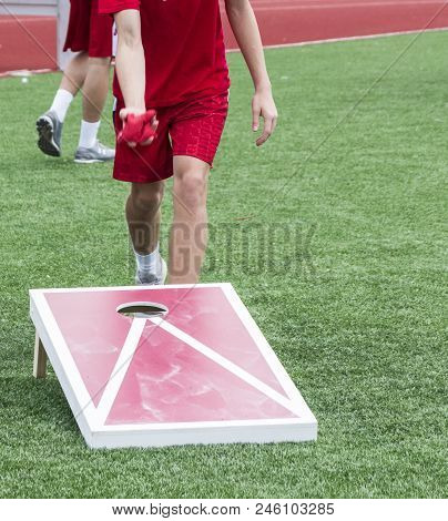 A Kid Is Throwing A Bean Bag During A Game Of Corn Hole On A Turf Field Outside.
