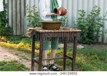 A Boy In A White Shirt And A Green Sweater, Holds A Clay Jug, Pours Water Out Of It Into A Copper Ba
