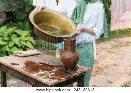 A Boy In A White Shirt And A Green Sweater, Holds A Clay Jug, Pours Water Out Of It Into A Copper Ba