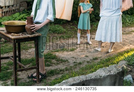 A Boy In A White Shirt And A Green Sweater, Holds A Clay Jug, Pours Water Out Of It Into A Copper Ba