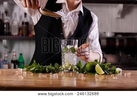 A Smiling Barman Puts Cubed Ice Into A Glass Of Refreshing Mojito, Decorated With Mint And Limes.