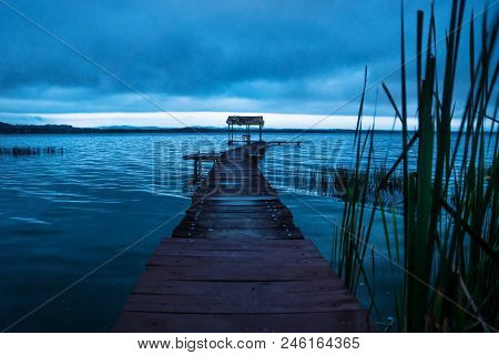 Pier With Hut Behind Moving Grass Along The Lake Shore Before Sunset In Blue, El Remate, Peten, Guat
