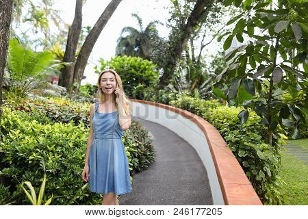 Young Caucasian Woman Talking By Smartphone Near Tropical Plants, Wearing Jeans Sundress. Concept Of