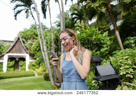 Young Pretty Girl Using Smartphone In Garden Near Trees, Wearing Jeans Sundress. Concept Of Summer V