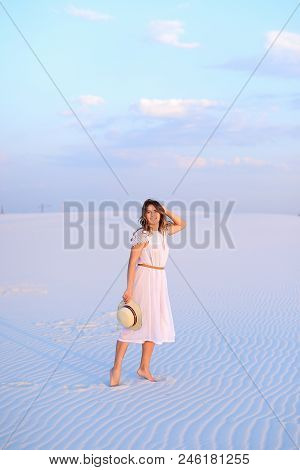 Young European Woman In White Dress Walking On Sand And Keeping Hat. Concept Of Photo Session In Des