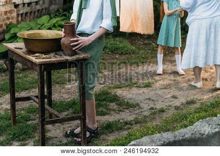 A Boy In A White Shirt And A Green Sweater, Holds A Clay Jug, Pours Water Out Of It Into A Copper Ba