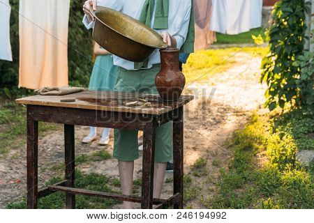 A Boy In A White Shirt And A Green Sweater, Holds A Clay Jug, Pours Water Out Of It Into A Copper Ba