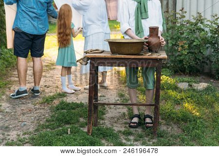 A Boy In A White Shirt And A Green Sweater, Holds A Clay Jug, Pours Water Out Of It Into A Copper Ba