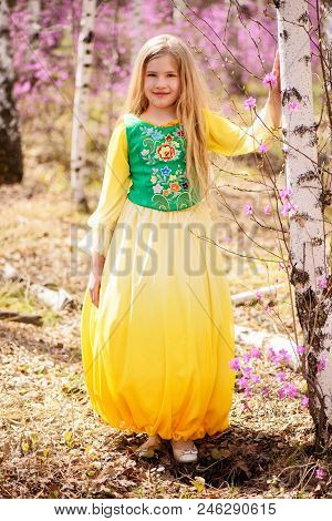 A Child With A Long Blond Hair Stands Among The Ledum And Birch In A Green Yellow Dress And Smiling