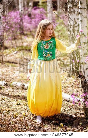 A Child With A Long Blond Hair Stands Among The Ledum And Birch In A Green Yellow Dress And Smiling