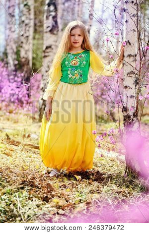 A Child With A Long Blond Hair Stands Among The Ledum And Birch In A Green Yellow Dress And Smiling