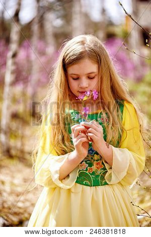 A Child With A Long Blond Hair Stands Among The Ledum And Birch In A Green Yellow Dress And Smiling
