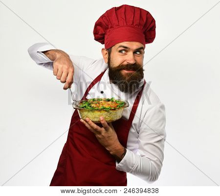 Cook With Tricky Face In Burgundy Uniform Holds Baked Dish. Man With Beard On White Background. Chef