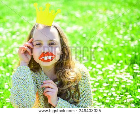 Girl Sits On Grass At Grassplot, Green Background. Child Posing With Cardboard Smiling Lips And Crow