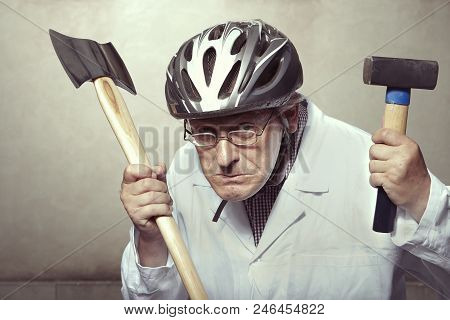 Older Paramedic Freak Man In Medical Coat And Protective Helmet Armed With Double Axe And Hammer