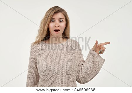 Portrait Of Astonished Attractive Young Woman With Index Finger Shows To The Side, Keeps Her Mouth W