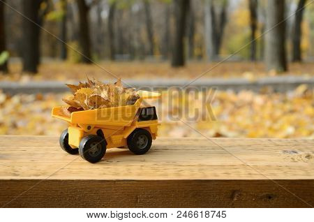 A Small Toy Yellow Truck Is Loaded With Yellow Fallen Leaves. The Car Stands On A Wooden Surface Aga