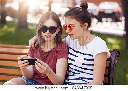 Best Friends Embrace While Sit On Bench In Summer Park, Watch Short Videos On Mobile Phone, Conncect