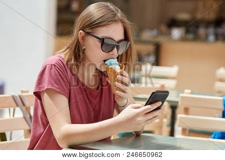 Pretty Young Female In Trendy Shades, Eats Ice Cream And Texts Messages On Smart Phone, Connceted To