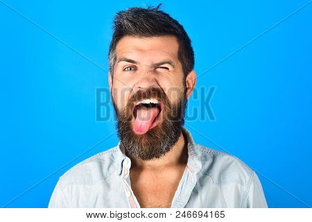 Bearded Man With Long Beard And Moustache Shows Tongue. Portrait Of Handsome Bearded Man On Blue Bac