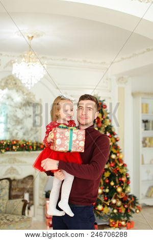 Young Father Keeping Little Daughter With Present, Christmas Tree In Background. Concept Of Gifts An