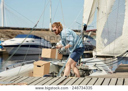 Young yachtsman holding box with provisions for weekend sea trip while working on yacht