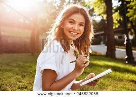 Portrait of pleased amazed woman 20s sitting on green grass in park with legs crossed during summer day and writing down notes with pen