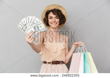 Portrait of an excited young woman in summer hat showing bunch of money banknotes while holding shopping bags isolated over gray background